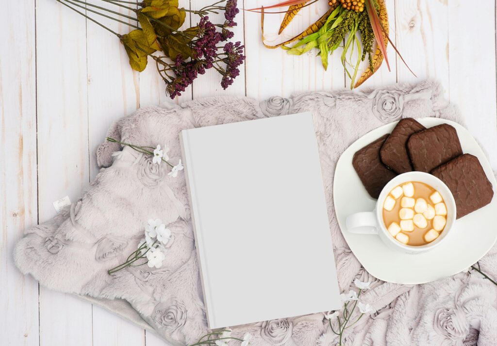 Warm and inviting flat lay with hot chocolate, cookies, book, and flowers on a soft blanket.