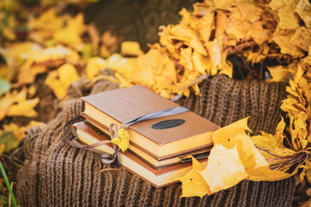 Stack of books with yellow fall leaves on a knitted blanket outdoors.