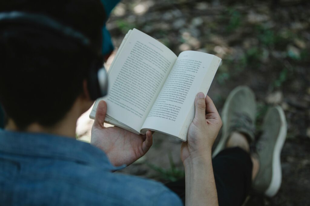A man enjoying reading a book and listening to music outdoors in a park setting.